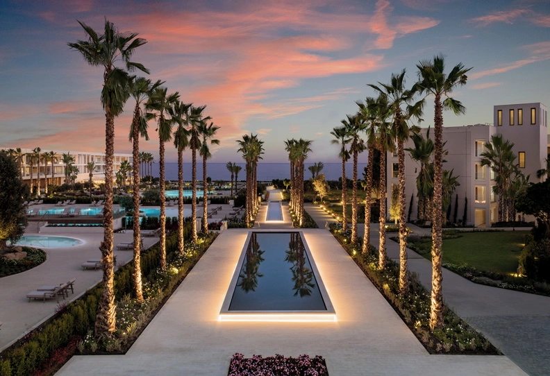 Palmengesäumter Pool des Ikos Resorts bei Sonnenuntergang mit beleuchtetem Gehweg