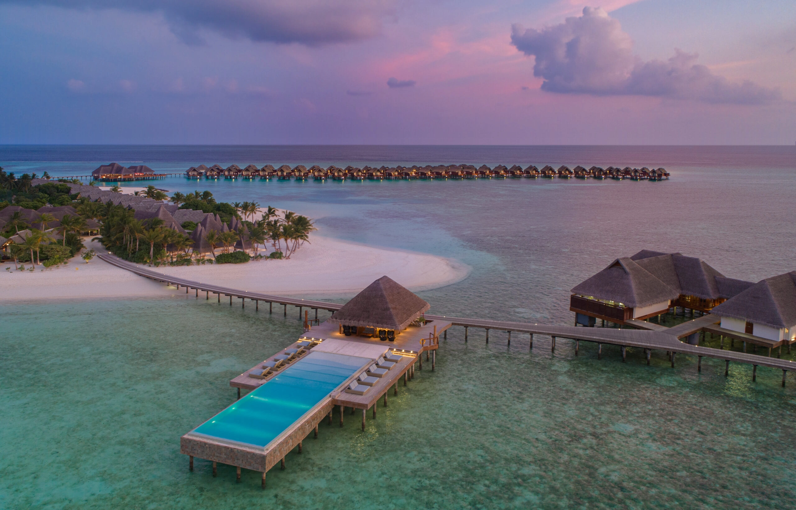 Infinity-Pool bei Sonnenuntergang mit Blick auf eine tropische Insel und Villen über dem Wasser auf den Malediven