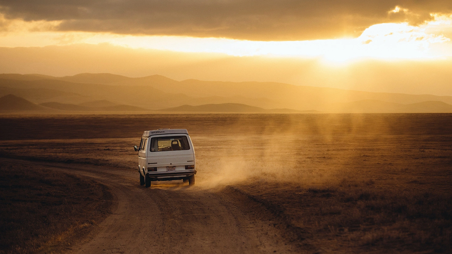 Van driving through desert landscape at sunset with dust trail and mountains in the background
